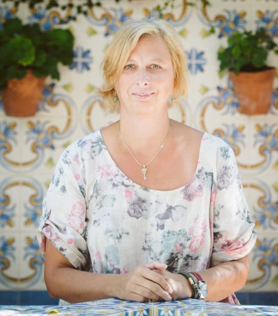 Woman smiling at the camera, sitting at a table with decorative tiles and potted plants in the background, promoting SLIMM weight loss supplement in a bright, inviting setting.